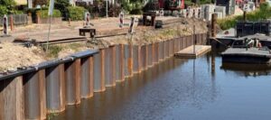 Construction work along a riverbank with a steel sheet-pile wall, digging, and construction barriers visible along the edge.