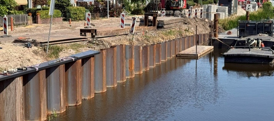 Construction work along a riverbank with a steel sheet-pile wall, digging, and construction barriers visible along the edge.