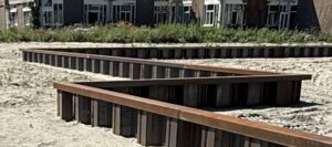 Rectangular wooden planters with rust-colored edges arranged in a stepped formation on a sandy lot; buildings visible in the background.