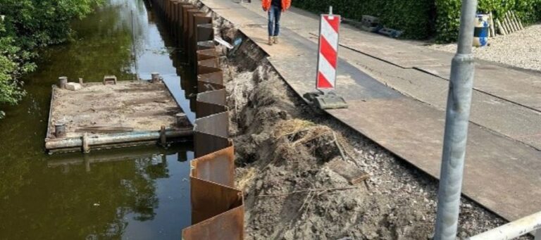 Construction work along a canal with a steel sheet pile wall and exposed soil; a worker stands on the adjacent sidewalk behind a striped safety barrier.