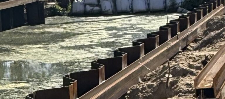 Construction site beside a murky body of water with metal sheet piles forming a barrier along the bank and exposed dirt on the right side of a trench edge.