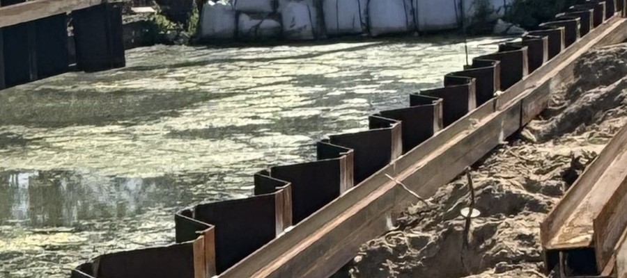 Construction site beside a murky body of water with metal sheet piles forming a barrier along the bank and exposed dirt on the right side of a trench edge.